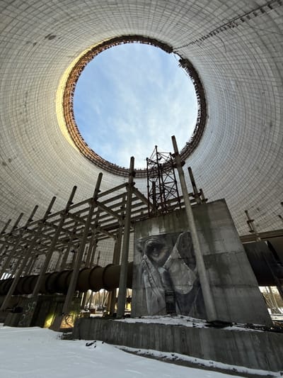 This image presents an upward-looking view from the interior of a massive, cylindrical concrete structure, characteristic of a cooling tower. The scene is set in what is likely the Chernobyl Exclusion Zone, specifically Pripyat, Ukraine.

The vast inner wall of the tower, constructed from light-colored, patterned concrete blocks, curves dramatically upwards, leading to a large circular opening at the top through which a clear blue sky with scattered white clouds is visible. On the right side of the frame, a complex network of concrete beams, metal girders, and wooden scaffolding-like structures ascends vertically, suggesting former industrial infrastructure or construction elements.

A prominent and striking feature is a large, grayscale mural painted on a flat concrete surface integrated into the right-hand structures. The artwork depicts a human face, with a solemn eye, part of a nose, and hands appearing to be covering or holding something close to the face, creating a sense of contemplation or sorrow. Below this mural, several large, dark pipes or conduits descend into the unseen lower parts of the tower.

The ground or base level visible in the bottom right corner is covered in snow, indicating cold weather conditions. Natural daylight illuminates the scene from above, highlighting the immense scale of the structure and the intricate details of the mural. The overall atmosphere evokes a powerful sense of abandonment, industrial decay, and the unexpected presence of human artistic expression within a historically significant and desolate landscape. No visible text is present in the image.