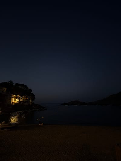 The image shows a nighttime scene in the town of Begur, Spain, likely near Sa Tuna or Aiguablava, coastal coves known for their picturesque setting and buildings. A cluster of buildings with illuminated windows are visible on a hillside to the left, overlooking the dark sea. Trees partially obscure the structures. In the water, several small boats are moored, some with lights reflecting on the surface. A sandy or pebbly beach occupies the foreground, with a single small boat pulled up onto it. Two individuals are seated on the beach, facing the water. The sky is a deep, dark blue, indicating it is night or very late dusk. The overall atmosphere is tranquil and serene.