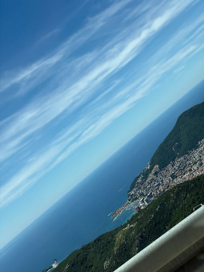 The image depicts an aerial view of the city of Stanišići, Montenegro, overlooking the Adriatic Sea. The perspective is angled, with the sky occupying the upper left portion of the frame and the sea and coastline extending diagonally downwards to the right. Wispy white clouds are scattered across the clear blue sky. The sea is a deep blue, with a few small boats visible. The coastline is lined with buildings, forming a dense urban area that cascades down the side of a lush green mountain. The mountain itself is covered in thick vegetation. In the foreground, a portion of a metallic guardrail is visible, suggesting the image was taken from a high vantage point, possibly a road or observation deck. The overall scene is one of a coastal city nestled between the sea and mountains.