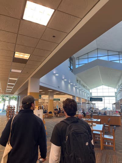 Two young people walk through a large library. The building has a high ceiling, with large windows allowing natural light. Bookshelves line the walls. Signage for "Information" and "Nonfiction" is visible. One individual wears a black backpack, while the other sports a baseball cap. The library appears modern, with sleek architecture and recessed lighting.
