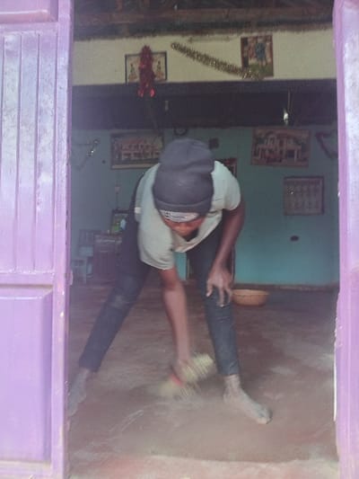 A person wearing a hat, light-colored shirt, and dark pants is crouched down, cleaning a wall with a brush. The wall is a rough, earthy color. The scene is viewed through a purple-painted structure, possibly a window or doorway. The interior of a room is visible in the background, decorated with wall hangings and a Christmas garland. A door stands open in the backdrop, partially obscured by the decor. The lighting appears somewhat dim, creating a slightly muted color palette in the room.