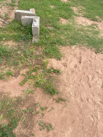 This is an outdoor, eye-level, medium shot of a patch of sandy ground with some grass and two concrete blocks. The scene is likely a natural or undeveloped area, possibly in Tavo, Nigeria, given the context. The lighting suggests it is daytime, likely sunny, with the sun casting subtle shadows.

The primary subjects are the two concrete blocks, which appear weathered and somewhat broken. They are positioned next to each other, with one block angled and leaning on the other. The larger block has some white paint remnants on its surface. The ground is covered in fine, reddish-brown sand, with clear imprints of what appear to be footprints. Patches of green grass and weeds are growing amongst the sand. The overall impression is one of an outdoor space that has seen some foot traffic, possibly a pathway or an informal gathering spot. There is no discernible activity or event taking place in the image; it appears to be a still-life captured in a natural setting. No text is clearly visible on the concrete blocks or elsewhere in the image. The overall mood is neutral and documentary, focusing on the textures and elements of the environment.