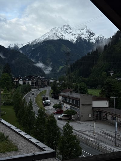 This is a medium-shot, vertical, eye-level, outdoor photograph of the village of Mayrhofen, Austria, on a cloudy day. In the foreground, a dark wooden railing is visible, with a gravel roof below it. A paved road curves through the scene, with several cars parked alongside it, including a white van, a dark SUV, and a red sedan. Buildings line the road, with a two-story structure featuring a dark roof and light-colored facade, and a larger building with a gray brick exterior and a covered entryway with glass doors. Trees and green grass are scattered throughout the scene. In the background, lush green mountains rise, with a prominent snow-capped peak dominating the skyline. A transmission tower and power lines are visible on the mountainside, with mist or low clouds partially obscuring the upper slopes. The overall atmosphere of the image is serene and a bit moody due to the overcast sky.