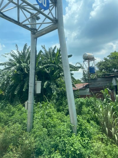 The image depicts a roadside scene with lush green vegetation, including banana trees and tall grass. A large metal support structure, likely for a sign, dominates the left side of the frame, with a blue circular sign with white lettering partially visible at the top. Several electrical conduits are attached to the support pole. To the right, a corrugated metal roof structure is visible, topped with a water heater and a blue plastic water tank on a metal frame. The sky is partly cloudy. There are no people visible in the image. The scene suggests a semi-rural or undeveloped area within Bình Xuân 2, Vietnam.