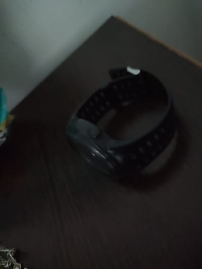 A black digital watch with a perforated band lies on a dark brown, wooden surface. The watch is positioned at an angle, with the face facing upwards. The surface appears to be a tabletop or desk. The background is a light gray wall, with a shadow cast from an unseen object. The lighting is dim, creating a somewhat blurry and low-contrast image.