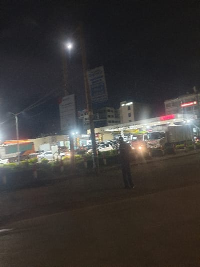 This media file captures a nighttime street scene in what appears to be an urban or semi-urban area, likely in Matathani ward, Kenya. The image is taken at night, with artificial lights illuminating the surroundings against a dark sky.

The scene features a street lined with multi-story buildings on the right side of the frame. These buildings display various illuminated signs and windows. Several vehicles, including cars and possibly motorbikes, are parked along the street. Headlights and taillights from some vehicles contribute to the bright light sources.

A single individual, dressed in dark clothing, is visible standing or walking on the paved area of the street. To the left, a prominent utility pole is seen, adorned with multiple signs. One sign has illegible blue text, and another below it has illegible white text on a dark background. A very bright, circular light source, possibly the moon or a powerful street lamp, is visible in the dark sky on the far left, exhibiting significant lens flare.

One of the buildings features a red illuminated sign with partially visible text, appearing to read "RU...". The overall atmosphere is dark with sharp contrasts from the numerous artificial light sources. No specific activities beyond general street presence and parked vehicles are discernible. The weather appears clear.