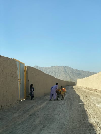 This image depicts a scene on a dusty, unpaved road in Mandisar, Afghanistan, under a clear, bright blue sky. A solitary individual, dressed in a light purple long-sleeved garment and pants, is actively pushing a yellow metal wheelbarrow along the dirt road, moving away from the viewer and slightly towards the right. The wheelbarrow contains at least two bright yellow jerry cans and possibly other unidentifiable items. To the left, a child in a multi-patterned long-sleeved dress stands stationary near a partially open, yellow-framed metal gate set into a tall, light-brown mud-brick wall. This wall runs parallel to the road on the left, curving slightly into the distance. Another similar mud-brick wall lines the right side of the road, also receding into the background. Distant, arid, light-colored mountains are visible under the clear sky. Strong shadows are cast on the road surface, indicating direct sunlight.
