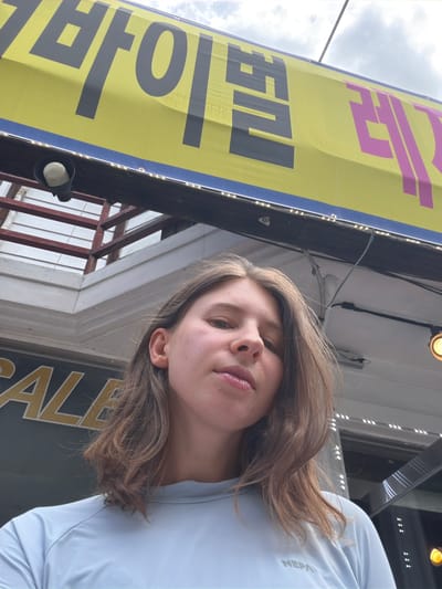 A young woman is pictured from a low angle, looking up and slightly to her left. She has long, light brown hair and is wearing a light blue t-shirt. Behind her, a large yellow banner with Korean text in black and pink is visible, as well as part of a building's exterior. The banner appears to be for a commercial establishment. A speaker is mounted on a dark beam above the woman. The overall scene suggests an outdoor setting, possibly a street or market area in Seo-myeon, South Korea.