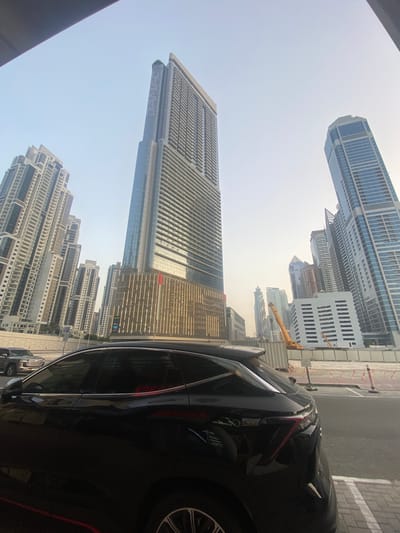 This photograph captures a partial view of the Business Bay area in the United Arab Emirates, focusing on the architecture and urban environment. The shot is taken from within or near a vehicle, with a portion of the black car visible in the right foreground. Multiple high-rise buildings dominate the skyline, including the Burj Khalifa, the tallest structure in the image, characterized by its distinctive tiered design and needle-like spire. To the left, other skyscrapers exhibit varying heights and architectural styles, with predominantly glass and steel facades. A yellow crane is visible amidst the buildings, suggesting ongoing construction or maintenance activities. The sky appears hazy, indicating potentially dusty conditions. The immediate foreground includes a portion of a road or sidewalk, indicating the image was captured at street level. The composition emphasizes the scale and density of the urban landscape, typical of Business Bay.