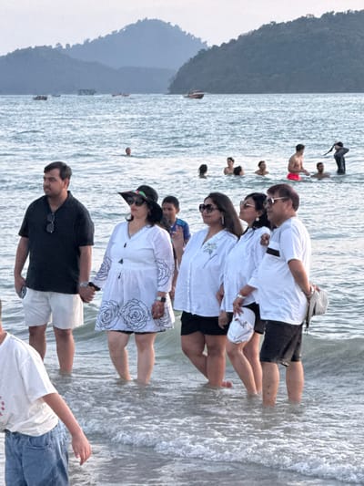 The image depicts a coastal scene at Cenang Beach, Malaysia. In the foreground and midground, a group of six adults and one child stands in the shallow ocean water, with waves reaching approximately mid-calf level. From left to right: a man in a black polo shirt and white shorts holds a smartphone and is holding hands with the woman next to him; he has sunglasses on his collar and a watch on his left wrist. Next, a woman wears a white long-sleeved tunic dress with black embroidered rose patterns, black shorts, a wide-brimmed black hat, and dark sunglasses; she also wears a watch on her left wrist. A young boy in a blue shirt is partially visible behind her. To their right, a woman in a white long-sleeved button-down shirt, black shorts, and dark sunglasses stands. Adjacent to her, another woman wears a white long-sleeved button-down shirt with a light green floral pattern, black shorts, and dark sunglasses, holding a white cap and a light-colored bag. The final individual in this primary group is an older man in a white polo shirt with a black stripe, dark shorts, and sunglasses, with a patterned bag slung over his shoulder. In the extreme lower left foreground, a child in a white t-shirt and blue denim shorts is partially visible, facing away from the camera.

In the mid-distance of the ocean, multiple other individuals are partially submerged, appearing to swim or stand in the water. One male figure is visible wearing red swimming shorts, and another is in a black, long-sleeved swimsuit or wetsuit. Several small boats are scattered across the water further in the background. The horizon is dominated by multiple large, vegetated landmasses or islands, suggesting a mountainous coastline, under an overcast sky.