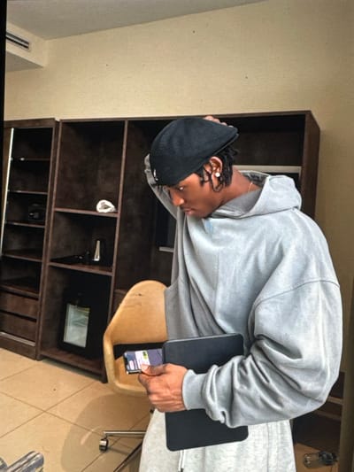 A young Black male is shown in a room, possibly a hotel or apartment, in Ifite Awka, Nigeria. He is wearing a grey oversized hoodie, grey sweatpants, and a black cap. His right hand is holding a black tablet, and his left hand is touching the back of his head, adjusting his cap. He is looking down and to his left, with his face partially obscured by his cap.
The room features a large, dark wooden bookshelf with multiple shelves displaying various items, including what appears to be a black speaker, a white paper bag, and a white rolled-up item. A tan chair with a metal base and wheels is positioned to the right of the bookshelf. The floor is tiled with a light beige color. In the background, there's a wall with a cream-colored finish and a horizontal air conditioning unit visible at the top left. The lighting suggests an indoor setting, with soft illumination casting shadows.