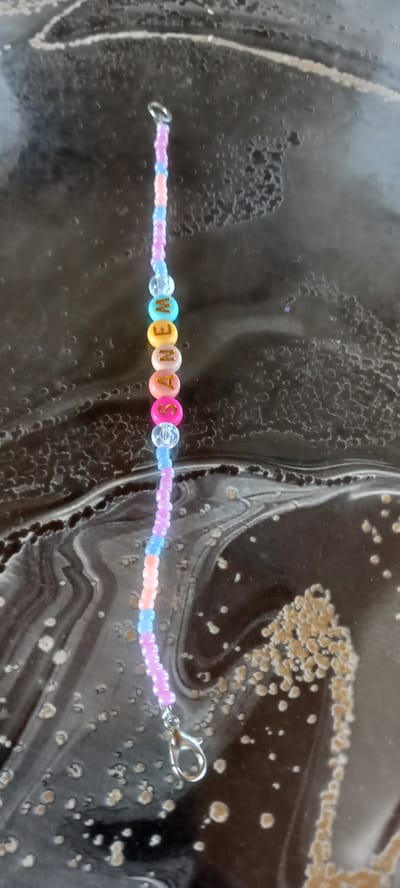 A beaded bracelet with the name "SASHA" spelled out in letter beads hangs vertically against a dark, textured surface. The beads are a mix of pastel colors, including pink, light blue, orange, and yellow, with some clear iridescent beads interspersed. The letters are gold and appear to be set into round, colored beads. A silver clasp is attached to the bottom of the bracelet. The surface beneath the bracelet appears to be a dark, glossy material with swirling patterns and lighter speckled areas. The location in the city of Iskra, Bulgaria, is not discernible from the image.