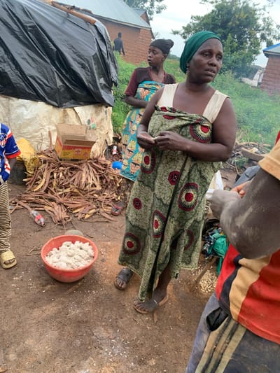A group of people are gathered outdoors in what appears to be a semi-rural or informal settlement. The ground is dirt, and there is debris scattered around, including dried plant material and a discarded plastic bottle. In the foreground, a woman wearing a green headscarf and a patterned dress stands looking to the right. She is holding a small white object in her hands. To her left, another woman in a patterned dress and a black headscarf is visible, looking to the left. A child wearing a blue, red, and white shirt is partially visible on the far left. In the center of the image, a red bowl contains a white powdery substance. Behind the people, a makeshift shelter is visible, covered with a large black tarp. The background consists of simple structures, trees, and vegetation, suggesting a warm climate. The overall scene depicts a moment of daily life and activity within a community. The location is identified as Buga da Gindi, Nigeria.