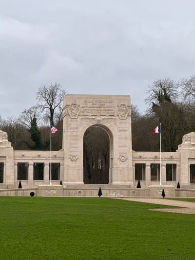 The image displays a grand stone memorial building under an overcast sky. The photograph is oriented vertically, but conceptually, the monument stands centrally with a green lawn to its right and a cloudy sky above.

The main structure is a large, light-colored stone archway, forming the focal point of the memorial. Above the arch, a prominent inscription is carved into the stone. The visible text reads:
"À LA MÉMOIRE
DES HÉROS
DE L'ESCADRILLE LAFAYETTE
MORTS POUR LA DÉFENSE
DU DROIT ET DE LA LIBERTÉ"
This translates to: "To the memory of the heroes of the Lafayette Escadrille who died for the defense of right and liberty."

The monument is adorned with intricate carvings and decorative motifs. Flanking the inscription, and elsewhere on the structure, are carved reliefs that appear to depict figures, possibly eagles, wings, or other symbolic elements associated with aviation and military honor. The architecture is grand and classical in style, with symmetrical sections extending from the central archway. These side sections feature rectangular recesses or openings, some appearing as windows or decorative niches.

Two flags are flying from flagpoles positioned in front of the monument. To the left side of the main archway (when viewed horizontally), a French flag (blue, white, and red vertical stripes) is visible. To the right side, an American flag (stars and stripes) flies. Both flags appear to be hanging relatively still, suggesting calm weather conditions.

In the foreground and to the right of the monument is a neatly manicured, vibrant green lawn. A lighter-colored pathway or gravel area runs along the edge of the lawn, leading towards the memorial.

Behind and around the monument, numerous trees, mostly deciduous, are visible. They are largely bare of leaves, indicating either winter or early spring. Some darker, possibly evergreen, foliage can also be seen. The sky overhead is uniformly light gray and overcast, suggesting a cloudy day.

The overall scene conveys a sense of solemnity and remembrance, characteristic of a war memorial or historical site.