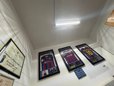 Three framed Barcelona football jerseys hang on a white wall. The first jersey features the Spotify logo, the second has the UNICEF logo, and the third is labeled "Ronaldinho 10". Below the jerseys is a display of small FIFA patches. To the left, a framed award with a blue ribbon is visible, along with part of another framed diploma. A long fluorescent light is mounted on the ceiling above.