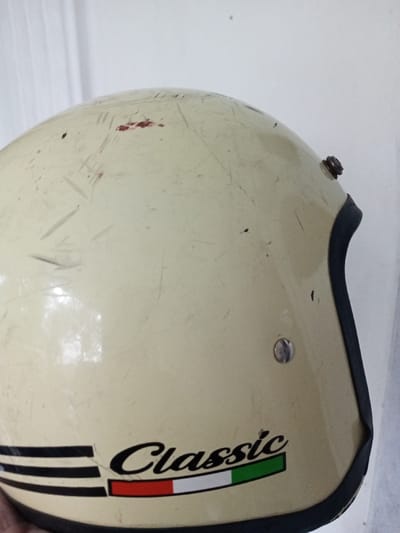 A well-used, light cream-colored motorcycle helmet is captured in the image. Prominent scratches and dark specks mark the helmet's surface. A "Classic" logo, complemented by the Italian flag's colors (red, white, and green), adorns the lower side. The helmet features a dark trim around the edge. A metal fastener is visible on the side, and another one sits near the top. Overall, the helmet shows signs of wear and tear.