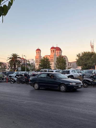 In Rethymno, Greece, this image depicts a parking lot filled with vehicles, including cars and motorcycles, with a church in the background. The church features red-domed roofs and a tall bell tower. The sky is a clear, pale blue, suggesting it is either early morning or late afternoon. Several green trees are visible, along with a tall palm tree. The parking lot pavement is dark asphalt. A dark colored sedan is in the foreground, with a person visible inside the driver's seat. The scene appears to be a common urban environment, likely in the city center given the presence of the church and the density of parked vehicles.