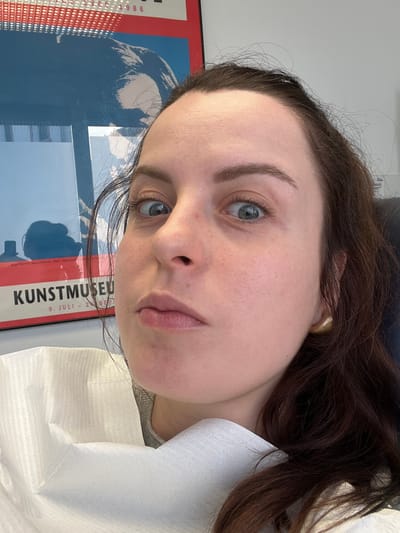 A female individual is depicted in a medium-shot, from the mid-chest upward, seated in what appears to be a dental or medical examination chair. She has fair skin, dark brown hair, and light-colored eyes, which are wide-open and directed towards the viewer. Her mouth is slightly pursed. A white, textured disposable dental bib is secured around her neck and upper torso. A gold-colored stud earring is visible on her right ear. In the background, a white wall holds a framed poster. The poster features a graphic design with red and blue sections, displaying the text "KUNSTMUSEUM," "9. JULI - 1986," and "Andy Warhol: Selbstbildnis, 1967. Siebdruck" in white lettering. Reflections, possibly from a window or light source, are present on the poster's surface, along with a dark silhouette. The scene takes place within a medical or dental clinic in Monsheim, Germany.