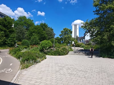 The image shows a bright, sunny day with a clear blue sky and scattered white clouds. In the foreground, a paved walkway made of light gray interlocking stones leads towards a set of stone stairs. A cyclist wearing a helmet and a light blue shirt rides on a paved path with bicycle symbols, which curves to the left. Adjacent to the path is a well-maintained garden with lush green bushes, flowering plants, and tall trees. In the background, a modern white tower with vertical green stripes and a clock face stands tall against the sky, identifiable as the church tower of St. Ulrich in Memmingen, Germany. To the right of the stairs, a woman dressed in black is walking up the steps, with another person further up the stairs. Buildings with light-colored facades are visible behind the trees.