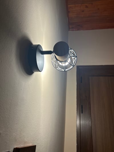 The image displays an indoor scene, likely a hallway or room, featuring a modern black wall sconce with a geometric metal shade illuminating a textured beige wall. To the right of the wall, a dark wooden door with vertical paneling is visible. Above, a wooden ceiling with visible planks runs across the top of the frame. No people are present in the image. The lighting suggests it is either nighttime or the ambient light is low, necessitating the use of the wall sconce. The image does not provide any specific indicators for the city of Sofronievo, Bulgaria.