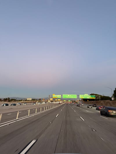 A long stretch of a highway with multiple lanes filled with cars during twilight. Overhead green signs are visible, directing traffic towards Irvine, Los Vegas, and San Diego. The sky is transitioning from blue to a soft pink-purple hue, creating a calming backdrop to the congested road. The perspective suggests the photo was taken from inside a vehicle traveling on the highway.