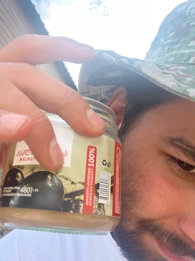 A close-up, selfie-style shot captures a person holding a jar of walnut paste. The person is wearing a camouflage bucket hat and their face is partially visible, focusing on their eye, cheek, and some facial hair. The jar label includes text in Georgian and English, indicating it is a "WALNUT" product and a "GEORGIAN PRODUCT 100%". A barcode is also visible on the label. The background appears to be an outdoor setting with a wooden structure and sky. The specific location is Arsha, Georgia.