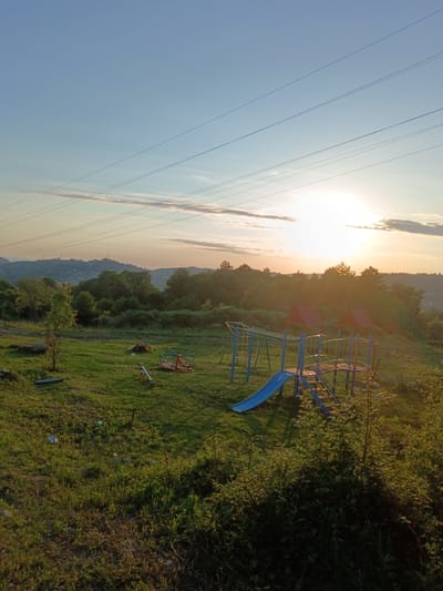 The image shows an outdoor playground scene at sunset in Chereshnya, Russia. The sun is low in the sky, casting a warm glow over the landscape. The playground equipment includes a blue slide with a metal frame, a merry-go-round, and a seesaw. The area is covered in green grass, with some scattered trees and bushes. In the background, rolling hills are visible, dotted with houses. Power lines stretch across the sky. The overall atmosphere is peaceful and serene, characteristic of a rural setting at dusk.