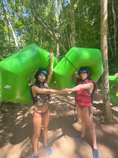 Two females are observed standing on a dirt path under a canopy of tall trees in a dense forest in Woods Town, Jamaica. Both individuals wear blue helmets and blue and black water shoes. The female on the left wears a camouflage-patterned life vest over a dark-colored bikini top and dark maroon bikini bottoms. She holds a large, bright green inflatable tube, with her right arm raised to grasp a black strap on the tube. Her left hand is extended, holding the right hand of the second female. The female on the right wears a red life vest over a dark green bikini. She also holds a large, bright green inflatable tube around her, with her left arm extended to hold the first female's hand. Both subjects face the camera directly, with the left female exhibiting a slightly puckered lip expression and the right female a neutral expression. Dappled sunlight illuminates sections of the path.