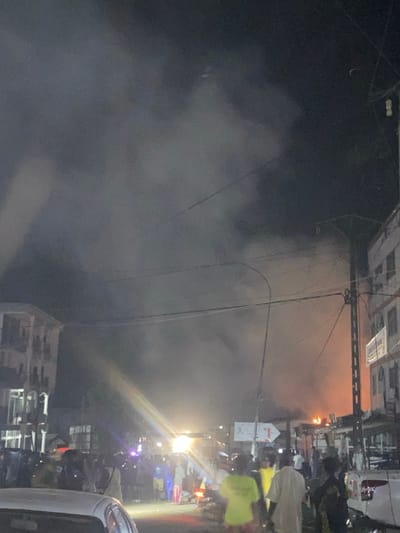 The image shows a street scene in the city of Limbe, Cameroon, at night. The location is near the coordinates 4.011, 9.216. A large amount of smoke billows from a fire on the far side of the street, illuminating the sky with an orange glow. There are numerous people on the street, standing and observing the fire. The majority of the people are wearing casual clothing, and many are holding cell phones. There are a few vehicles parked on the street, including a white sedan in the foreground, and a blue truck on the right side of the frame. Some street lights are lit, and multiple power lines crisscross the street. There is a building on the left side of the image, and another on the right. A sign with an arrow pointing left is visible on the right side of the street. A bright beam of light from a vehicle appears to be shining through the smoke. The fire appears to be in a building or group of buildings beyond the street.