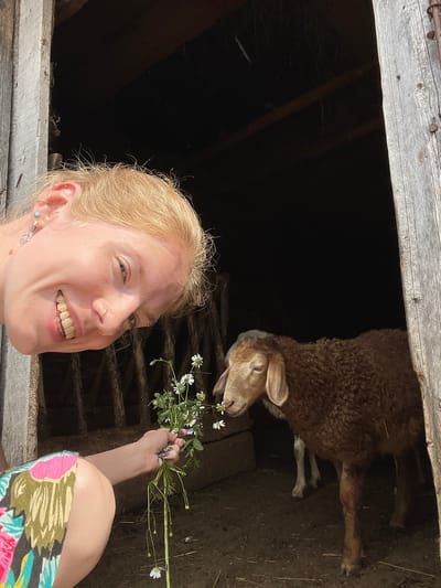 A young woman with blonde hair is taking a selfie with a sheep inside a rustic wooden structure. She is smiling broadly at the camera and holding a bouquet of small white wildflowers. The sheep, with its brown, curly wool, is reaching out towards the flowers, appearing to sniff them. The setting is dimly lit, with visible wooden beams and dark interior spaces suggesting a barn or shed. The woman is wearing a colorful patterned skirt and has several earrings and a nose ring. The time of day appears to be sunny, as evidenced by the bright lighting on the woman's face and the flowers. The overall mood is joyful and playful, capturing a moment of connection between the woman and the animal. The location context is Сям-Можга, Russia. There is no visible text in the image.