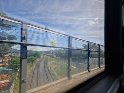 The image is taken from the interior of a moving vehicle, looking out a window. The window is a glass barrier with horizontal bars, offering a view of train tracks and the surrounding environment. In the foreground, a textured concrete surface is visible below the window.
The train tracks curve to the left, with ballast and sleepers clearly discernible. Further along the tracks, a white train is moving away from the viewer. The landscape beyond the tracks includes green trees and some industrial structures, including what appears to be a storage area with several freight containers. The sky is blue with wispy white clouds and contrails.
There are reflections on the glass, showing the sky and some trees. A yellow triangular warning sign is visible on the barrier. The overall scene suggests a journey through an urban or semi-urban area in Germany, specifically Walldorf, given the context provided. There are no people visible in the image.