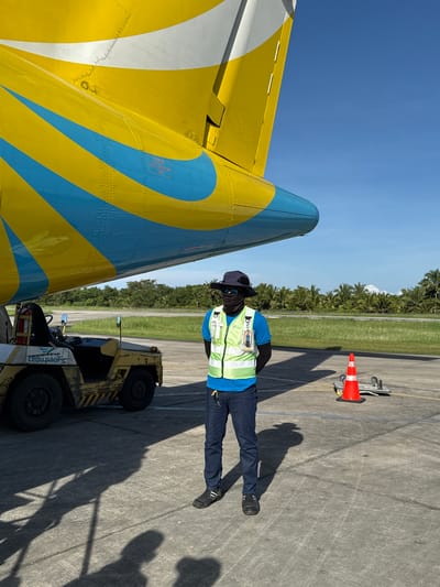 The image depicts an airport tarmac in Del Carmen, Philippines. A male airport worker is standing, wearing a bright green vest, dark pants, a blue shirt, a black face covering, sunglasses, and a hat. He is standing in front of a small yellow tow vehicle with black tires. Adjacent to him on the left is a section of an airplane, its tail painted in bright yellow, light blue, and white. Behind the worker is a grassy area, followed by a dense row of palm trees. A red and white safety cone is near the trees. The sky is clear and blue. The ground is concrete.