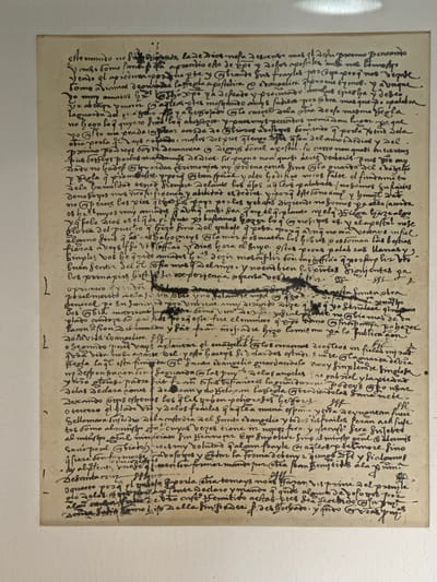 The image displays a single, aged sheet of paper, densely covered with handwritten text in dark brown or black ink. The paper has an off-white or cream color, indicating its historical nature, and shows signs of age such as slight discoloration, minor creasing, and several darker smudges or stains, particularly in the central and lower-right portions.
The text is written in a consistent, elaborate cursive script, characteristic of historical manuscripts, with varied line thickness and flourishes. It occupies the entire surface of the paper, from margin to margin. There are no depicted subjects, people, or objects other than the document itself; the image focuses entirely on the written content. The activity implied is the act of historical record-keeping or writing.
Notable details include the uniform style of the handwriting, suggesting a single scribe, and the evident age of the document. Throughout the text, small circles are drawn around certain letters or words, possibly serving as markers or references. At the bottom center of the page, the numeral '1' is visible in a modern font, likely a later addition for pagination or cataloging, distinct from the original historical script. The visible words suggest the language is Spanish, consistent with the historical context of Tepotzotlán, Mexico. The document appears to be a significant historical manuscript.
