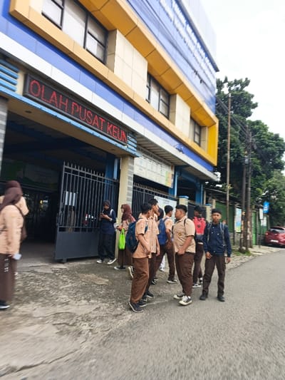 A group of students in brown and beige uniforms gathers outside a building with a blue and yellow facade. The building has a sign that reads "OLAH PUSAT KEUN." Some students stand near an open gate, while others are closer to the street. A red car is visible in the background, parked near trees and a street sign. The sky is overcast.