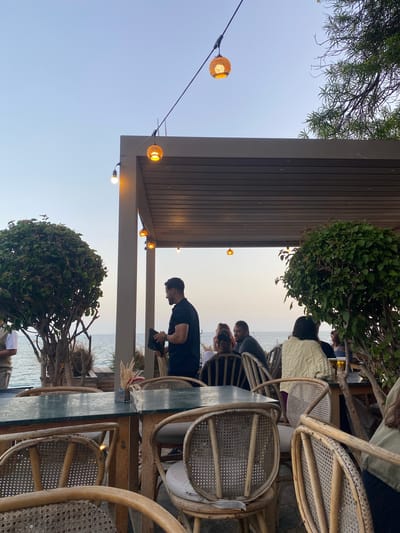A group of people are seated at tables in an outdoor restaurant with a view of the sea. A man in a black shirt is standing and holding a black object. There are string lights overhead and trees surrounding the dining area. The scene appears to be in Taghazout, Morocco, given the architecture and the presence of the sea. The lighting suggests it is either dawn or dusk.