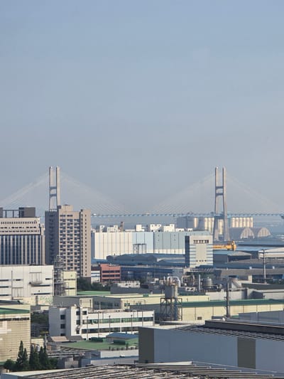 This media file displays a high-angle, sideways view of an industrial cityscape. The scene is dominated by a variety of buildings, including warehouses, factories, and office buildings, with a prominent suspension bridge stretching across the background. The weather appears hazy, suggesting a light fog or smog, and the time of day is likely daytime, given the illumination.
The main subjects are the urban industrial structures and the bridge. Among the buildings, some are tall and modern with numerous windows, while others are lower and appear to be functional industrial facilities. The suspension bridge, a significant object in the scene, features two tall concrete towers supporting a network of cables that fan out to hold the bridge deck. A yellow crane is visible near the bridge, suggesting ongoing construction or industrial activity.
There is some visible text on the structures. On some of the large cylindrical tanks in the background, Korean text is present, though it is partially obscured and difficult to read fully. A yellow crane has the word "JOYANG" and what appears to be "HOIST" written on it. On the lower right portion of the image, on a white building, partially visible Korean text can be seen, including "GINDOO FINE CHEA".
The overall impression is one of a busy industrial hub, possibly a port area given the presence of the bridge and the scale of the facilities. The hazy atmosphere lends a somewhat muted tone to the scene. No people are directly visible in the media file.