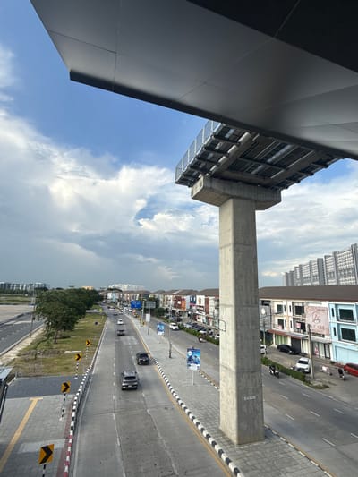 An elevated perspective captures a street scene in Pak Kret City Municipality, Thailand, under a partly cloudy sky. A large, dark gray overhang partially obscures the top of the frame, supported by a substantial concrete pillar that stands prominently in the mid-right of the image. The pillar is labeled with the text "MT02-P01".

Below the overhang, a multi-lane road stretches into the distance. On the right side of the road, several buildings with shops and businesses are visible, featuring colorful signage. A white car and a motorcycle are parked in front of one of these buildings. Further down the road, more cars and motorcycles are in motion, indicating moderate traffic.

On the left side of the road, a green median strip separates the lanes from a sidewalk area. This sidewalk features yellow tactile paving. Several yellow and black chevron signs are posted along the roadside, indicating curves or hazards. In the background, a row of trees lines the median, and beyond them, apartment buildings rise against the sky. The overall impression is of a functional urban environment on a day with variable weather.