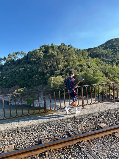 A young person, wearing a cap, backpack, and athletic clothing, walks along a railway bridge in Générargues, France, holding a rock. The bridge has a rusted metal railing and a stone walkway bordering the railway tracks. Below the bridge, a river flows, bordered by rocks and a sandy bank. The background features a lush, tree-covered hillside under a clear blue sky. The sunlight casts long shadows, indicating it is late afternoon.