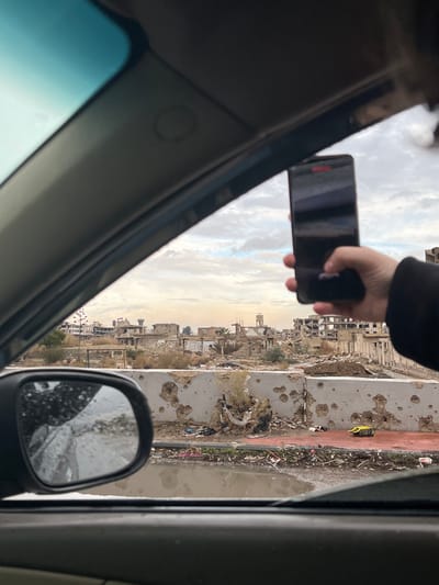 The image captures a perspective from inside a vehicle, looking out the passenger-side window at an urban landscape identified as Al-Qaboun Municipality, Syria.

In the foreground, the vehicle's dark gray interior elements, including the A-pillar and roof lining, are visible. A right hand holds a black smartphone vertically, obscuring the upper-right portion of the view. The phone's screen displays reflections of the scene, and a red banner-like notification is partially visible at the top. The person's thumb rests on the lower part of the screen. The vehicle's side mirror is visible in the lower-left, reflecting a wet road surface and debris.

The midground features a concrete barrier, heavily marked with numerous pockmarks and craters consistent with bullet or shrapnel impacts. Below this barrier, a narrow strip of ground is covered with sparse, dry vegetation and accumulated debris, including what appears to be discarded plastic and fabric. Adjacent to this strip is a muddy ditch or canal containing standing water, also littered with trash.

The background reveals an extensively damaged urban environment under an overcast sky with scattered clouds. Numerous buildings appear severely dilapidated, reduced to skeletal structures with missing roofs, walls, and windows. Visible structures include multi-story residential or commercial buildings in various states of disrepair. A prominent water tower stands on the right horizon, and a structure resembling a minaret or bell tower is visible slightly left of center. The landscape is desolate, with minimal intact vegetation, primarily withered trees and shrubs scattered among the rubble. The overall scene indicates widespread destruction.