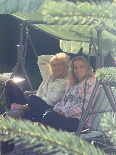 In this photo, two women are sitting on a swing set outdoors. The woman in the foreground is wearing a light-colored, fuzzy sweater and dark pants. She is leaning back, with one arm resting on her head, and her legs are crossed. Her expression appears relaxed. The woman behind her is wearing a pink plaid shirt with a graphic print. She is smiling and looking towards the camera. Sunlight filters through the branches of a tree, casting dappled light on the scene. The background is out of focus, but appears to be a grassy area. The time of day seems to be late afternoon or early evening, given the warm sunlight. The overall mood is one of contentment and casual relaxation. The setting suggests a backyard or garden in Kerro, Russia. There is no visible text in the image.