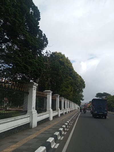 The image depicts a roadside scene in Thotagamuwa, Sri Lanka, under an overcast sky. The vantage point is from the road, looking forward.
On the left, a long, ornate fence stretches into the distance. This fence is composed of regularly spaced white concrete pillars with decorative capitals, connected by dark, wrought-iron sections topped with golden spikes. Behind the fence, a dense line of tall, dark green coniferous trees, possibly cypress, forms a prominent natural barrier, with their foliage extending high into the sky.
Adjacent to the fence is a paved sidewalk, constructed from grey rectangular tiles, featuring a yellow tactile paving strip running parallel to the curb. A black and white striped curb separates the sidewalk from the two-lane asphalt road.
On the right side of the image, the dark grey asphalt road is visible. A dark blue box truck is positioned in the right lane, moving away from the viewer, with its rear facing the camera. Behind and to the left of the truck, at least two motorcycles are present, each operated by a rider, also traveling in the same direction. Additional vehicles are indistinctly visible further down the road, suggesting ongoing traffic.
The sky is predominantly light grey and overcast with dense cloud cover, though some lighter patches are discernible. In the far distance, beyond the road and surrounding vegetation, elements of a town or city, including buildings and more trees, are visible beneath the cloudy horizon. The overall lighting is diffused, consistent with an overcast day.