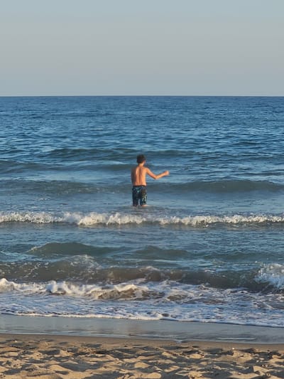 This media file depicts a young boy in mid-air, appearing to jump or dive into the ocean. He is wearing patterned swim trunks and his body is angled downwards, suggesting a dynamic action. The boy is positioned in the water, with waves around him. To the right of the water is a sandy beach. The sky is a clear, light blue, indicating fair weather. The scene appears to be set during the daytime, possibly late afternoon given the lighting on the sand. The overall impression is one of youthful energy and enjoyment of a beach environment. The location is identified as Castelldefels, Spain. There is no visible text in the media file.