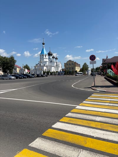 This image shows a street scene in Pastavy, Belarus. In the background, a white Orthodox church with blue domes and gold accents is visible. Several parked cars line the street to the left of the church. In the foreground, a pedestrian crossing with yellow and white stripes is painted on the asphalt road. A traffic sign with a red circle and a blue cross inside is posted on a pole to the right of the road. The sky is blue with scattered clouds.