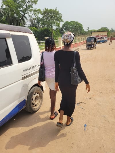 A daylight photograph captures an outdoor scene on an unpaved road in Candelaria, Colombia. Two adult females walk away from the camera, moving towards the right side of the frame. The woman on the right wears a long-sleeved black dress, a colorful patterned headscarf, and black slides, carrying a black textured handbag on her right shoulder. The woman to her left has dark braided hair with lighter ends, wears a light pink top, white shorts, and black and red slides, and carries a black handbag on her left shoulder. Both cast distinct shadows on the light brown dirt ground, which contains a small, metallic spoon-like object in the lower right foreground.

To the immediate left of the women, the rear portion of a white van is visible, featuring dark tinted windows, a blue stripe along the lower body panel, and a worn rear tire. Faint, illegible text is present on the van's side. In the mid-ground, a bridge structure with cream-colored pillars and a red top rail crosses the scene. Beyond the bridge, a black three-wheeled auto-rickshaw (tuk-tuk) and a light blue and white small van are visible, both moving away from the viewer. Further in the background, a person in a brown uniform stands near a structure. The background is characterized by lush green tropical vegetation under a bright, clear sky.