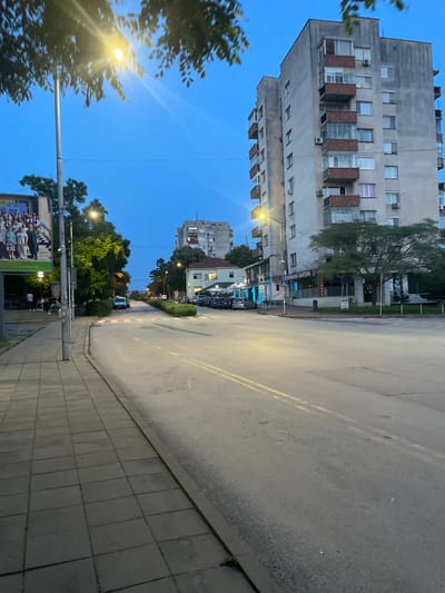 The image depicts a street scene in Chirpan, Bulgaria, during the early evening or dusk. The sky overhead is a deep blue, indicative of twilight, with a bright streetlamp in the upper left foreground emitting a strong yellow light and lens flare, partially obscured by dark green tree foliage. Another streetlamp is visible further down the street on the left.
On the right side of the image, a large, multi-story concrete apartment building dominates the midground. The building exhibits a weathered, grey facade with numerous windows and several enclosed balconies, some with visible air conditioning units. Its design suggests a functional, possibly socialist-era architectural style.
The foreground features a grey tiled sidewalk on the left, leading to an asphalt road with a dark grey curb. The road has faint double yellow lines running down its center. Further down the street on the right, smaller, single-story buildings are visible, one of which has a neon blue sign with Cyrillic text reading "НОТАРИАЛНА КАНТОРА" (Notary Office). Multiple cars are parked along both sides of the street in the mid-distance. A white sedan is parked on the left side, further down the street.
On the far left, a large billboard-like structure displays an image of a group of people, possibly a school or event photograph. Beneath this billboard, a group of approximately five to seven individuals is gathered on the sidewalk, some seated, some standing. Sparse green trees and manicured hedges line portions of the street and sidewalk. The overall lighting suggests artificial illumination from streetlights is becoming dominant as natural light fades.