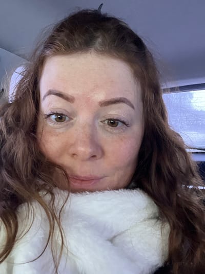 The scene is located in Semenovo, Russia. The image displays a close-up frontal view of an adult female. She has reddish-brown, wavy hair, with a dark hair clip visible securing hair at the top of her head. Her skin is fair with visible freckles, and she has brown eyes and well-defined eyebrows. Her lips are closed, forming a slight smile. She is enveloped in a thick, white, plush scarf or collar that covers her neck and lower face, partially obscuring a dark garment beneath. The background on the left is a uniformly light, blurred surface, possibly an interior vehicle component. On the right, through a blurred window, faint outlines of what appear to be frosty trees or vegetation are visible under diffuse, cool lighting. The individual is looking directly towards the camera, suggesting a self-portrait.