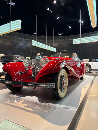 The image shows a red 1936 Mercedes-Benz 500K Spezial-Roadster on display in a museum. The car is prominently featured in the foreground, with its sleek, classic lines and chrome accents catching the light. In the background, other vintage cars are visible, along with display lighting and dark walls. A descriptive plaque at the bottom left of the image identifies the car and its specifications. The overall setting appears to be a well-lit exhibition hall, suggesting a museum dedicated to automotive history. This particular car and its display are likely located at the Mercedes-Benz Museum in Stuttgart, Germany, given the subject matter and the museum's reputation.