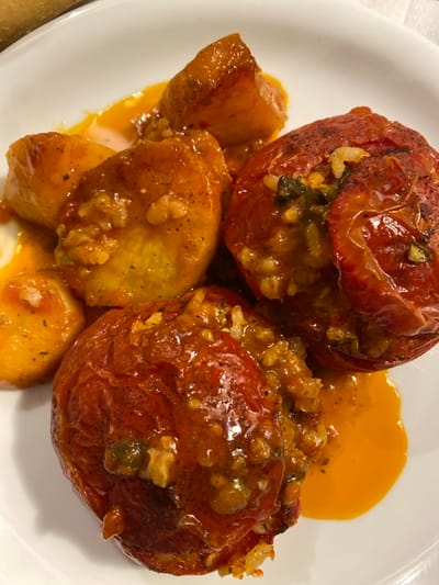 A close-up view presents a meal served on a white, round plate. The primary subjects are two whole, red tomatoes visibly stuffed with a mixture of rice and green herbs, appearing cooked and slightly softened. Several irregularly shaped, roasted potato pieces, exhibiting a light yellow to orange-brown color, are arranged around the stuffed tomatoes. A vibrant orange-red sauce coats parts of both the tomatoes and potatoes, pooling on the white plate. Dark specks, consistent with ground pepper or dried herbs, are scattered across the food items. No people or specific environmental details beyond the plate are discernible. The scene is located in Megalochori, Greece.