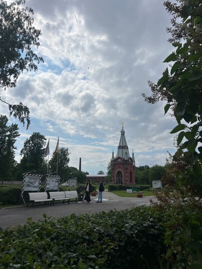 In the city of Kronstadt, Russia, an outdoor scene unfolds under a cloudy sky. A red, ornate chapel with a tall, multi-colored spire topped with a golden cross stands prominently in the background, framed by trees. In the middle ground, three individuals are present: two are standing near a white bench, and one is bending down. Two flags, one red and one yellow with symbols, are planted near them. Educational displays on black and white stands are also visible. A paved path leads through the area, bordered by green bushes and trees. In the distance, a tall brick structure, possibly a chimney, and a building with a red roof are discernible.