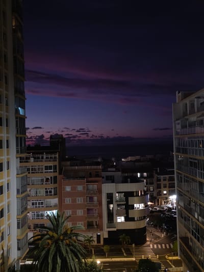 This media file depicts a twilight scene in an urban environment, likely Puerto de la Cruz, Spain. The dominant subjects are a series of multi-story buildings, characterized by modern architecture with glass facades and balconies. Some buildings feature prominent curved designs. A large palm tree with visible fronds and some yellowing, possibly fruiting, structures stands to the right of the frame, adding a tropical element to the cityscape.

The sky displays the deep purples and subtle pinks of dusk, with a few scattered clouds. Lights are visible in many of the building windows, indicating that it is evening. Below the buildings, a street is visible with crosswalks and parked vehicles, suggesting a residential or commercial area. The activity appears to be the quiet transition from day to night in a city, with no specific event or action prominently featured.

Notable details include the time of day, which is clearly twilight or early evening, the urban setting with its architecture and streetscape, and the presence of the palm tree hinting at a warmer climate. The lighting conditions suggest the ambient light from the setting sun mixing with artificial lights from the buildings and street. There is no discernible text within the image. The overall mood conveyed is one of calm and observation of a city at dusk.